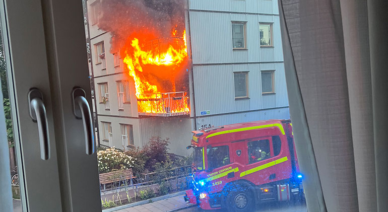 Full utvecklad brand i två balkonger i ett lägenhetshus, röken döljer ytterligare en balkong ovanför. En brandbil står på gatan. Bilden tagen genom ett fönster i ett hus mittemot.