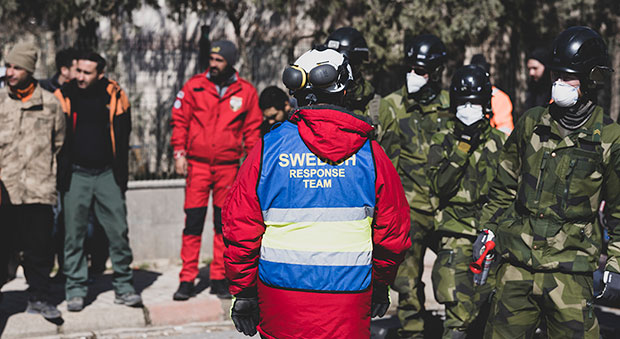 Delar av det svenska teamet, bestående av personer i militärkläder och en MSB-anställd med ryggen mot kameran. Den sistnämnda har en röd jacka med en blå väst över där det står Swedish response team.