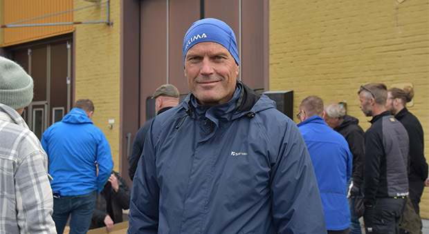 Göran Grudén, klädd i blå vindjacka och blå mössa, står framför en gul tegelvägg. Mellan honom och väggen ser man ryggen på en grupp andra människor.