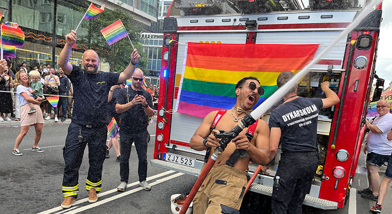 En brfandman sprutar vatten med en slang på Pridefestivalen. Bakom honom är en brandbil och på gatan ses flera brandmän som viftar med regnbågsflaggor.