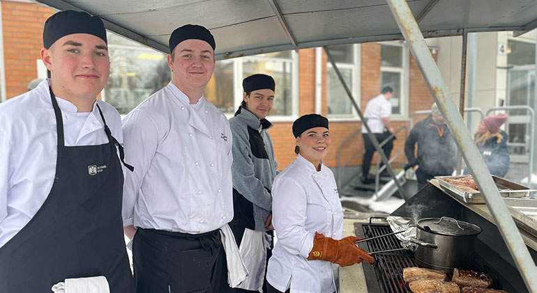Fyra gymnasieelever i kockkläder grillar kött utomhus.