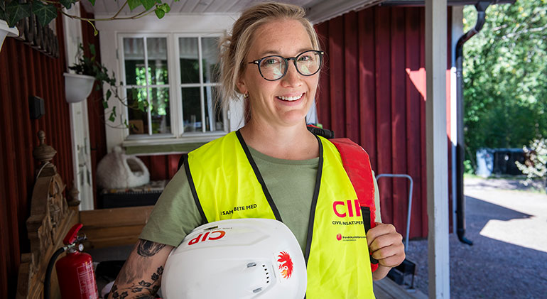 Hanna Norin står utanför sitt röda hus med gul CIP-väst på och en vit CIP-hjälm h handen.