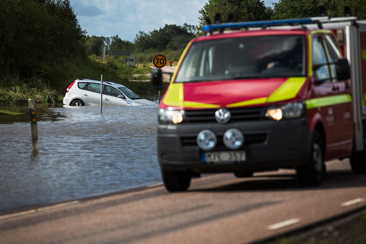 Bil från räddningstjänsten i förgrunden. I bakgrunden har en personbil hamnat i vattnet vid en översvämning.