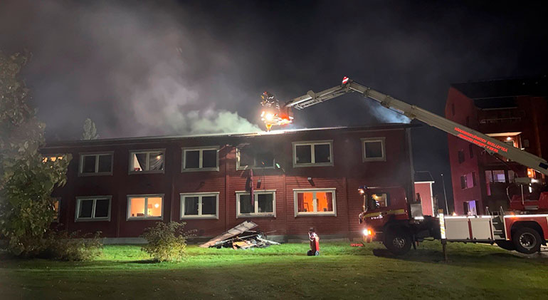 Ett rött hus brinner, det kommer rök från taket och man ser lite av en brandbil till höger i bilden.