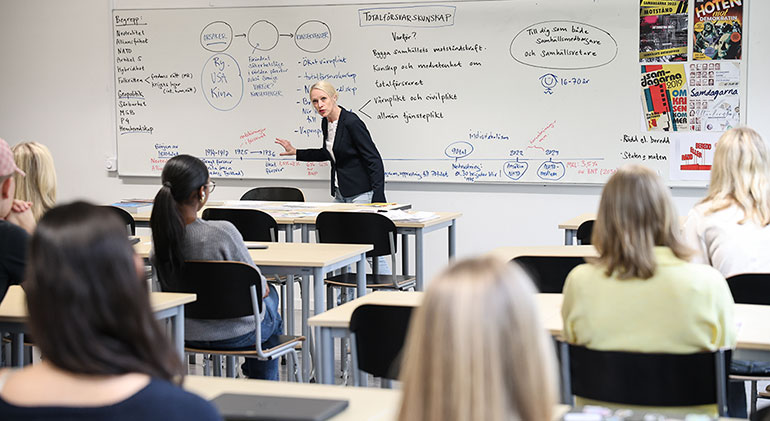 Läraren Karolina Kalat står vid whiteboard-tavlan i klassrummet och undervisar. Man ser ryggen på eleverna.