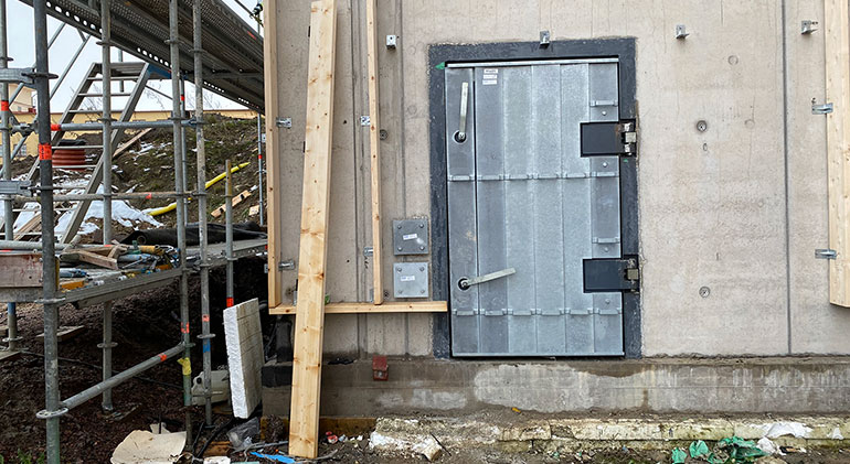 Ett skyddsrum som håller på att byggas. En stabil dörr är insatt och man ser byggnadsställningar bredvid.