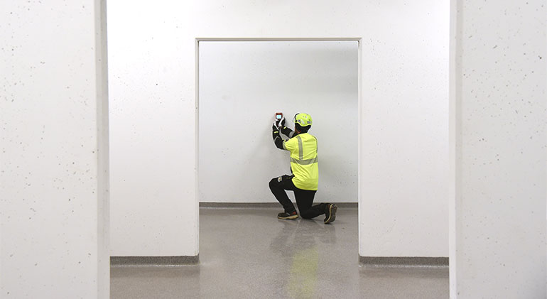 Lars Gråbergs, iförd skyddshjälm och varselväst, står på ett knä och mäter betongväggens täckskickt med en täckskiktsmätare.